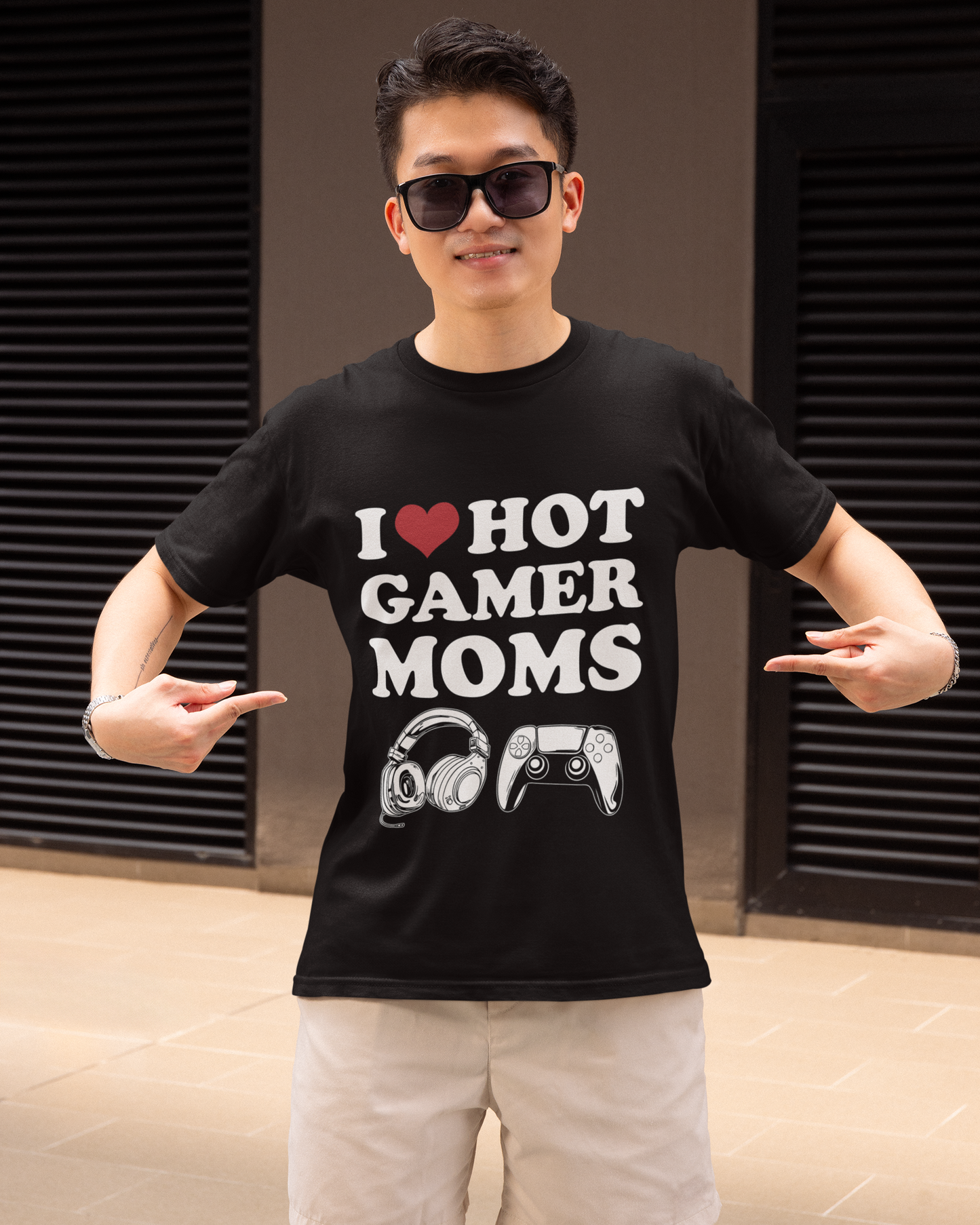 A man wearing black sunglasses and a black graphic t-shirt that reads 'I [heart] HOT GAMER MOMS' with a headset and controller graphic, paired with beige shorts, posing in front of a modern building with dark shutters.