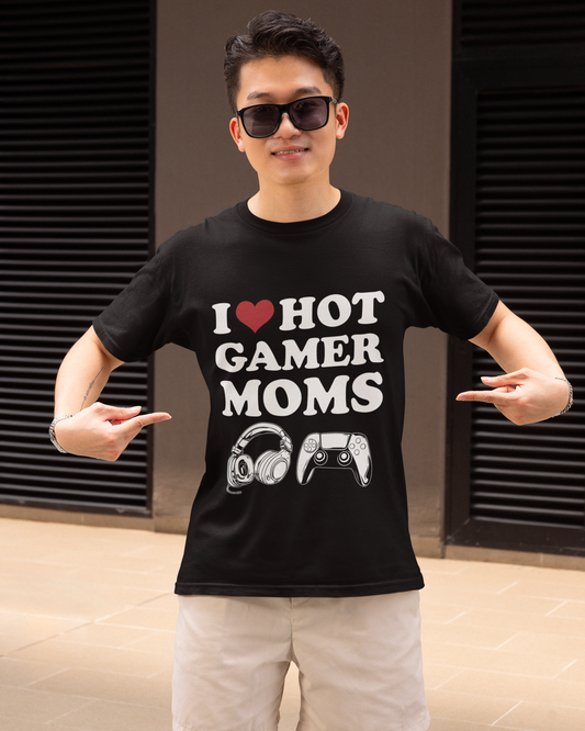 A man wearing black sunglasses and a black graphic t-shirt that reads 'I [heart] HOT GAMER MOMS' with a headset and controller graphic, paired with beige shorts, posing in front of a modern building with dark shutters.