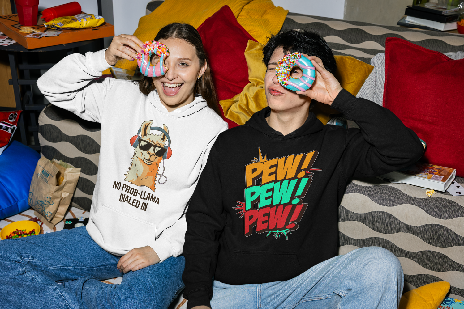 Two people sitting on a couch holding donut-shaped objects, one wearing a white hoodie with llama graphic and text, the other wearing a black hoodie with 'PEW! PEW! PEW!' text and graphics.