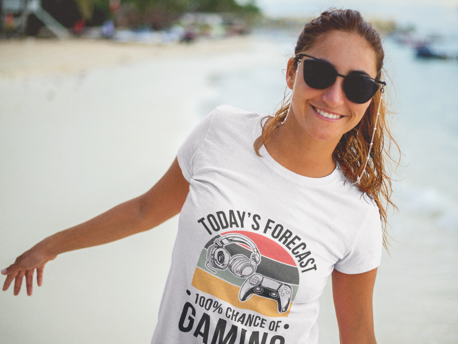 A woman wearing a white graphic t-shirt with a gaming design and sunglasses, standing on a sandy beach with the ocean in the background.