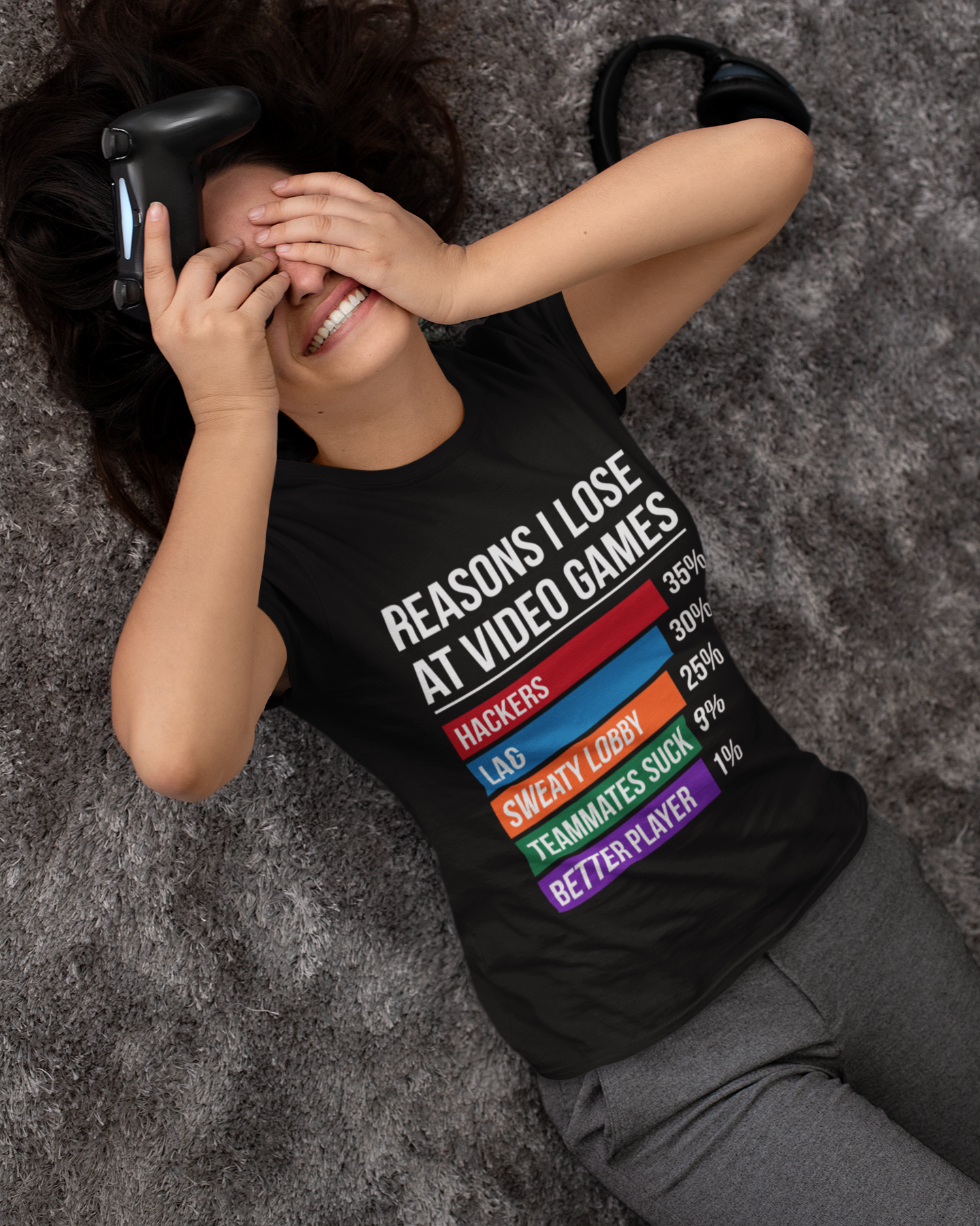 A woman lying on a gray textured rug wearing a black graphic t-shirt with a colorful list of reasons to lose at video games, along with gray sweatpants and black headphones.