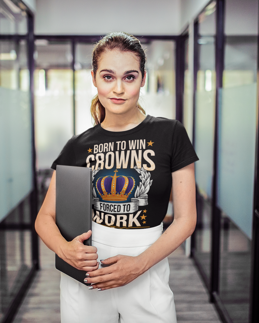 A woman standing in a modern office hallway wearing a black graphic t-shirt with a crown and the text 'BORN TO WIN CROWNS FORCED TO WORK' in beige and gold, paired with white pants.